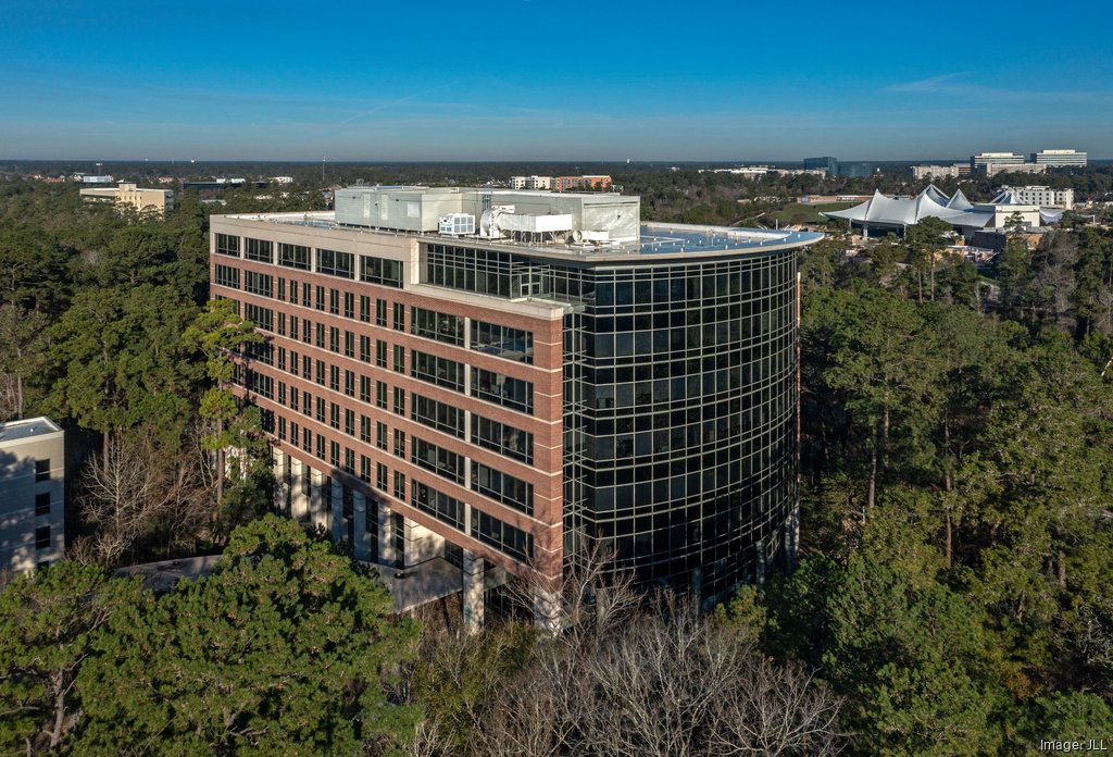 Multi-story modern office building in a wooded setting under a blue cloudless sky with other commercial properties in the distance.