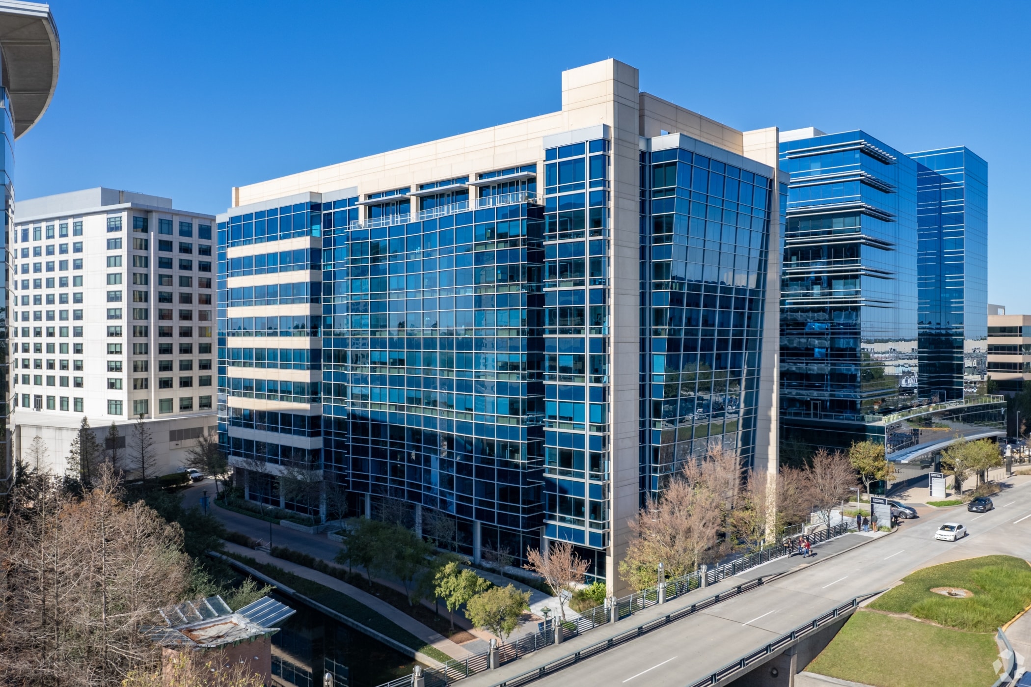 Modern multi-story office building with large glass windows in an urban setting surrounded by trees with other modern buildings around it.